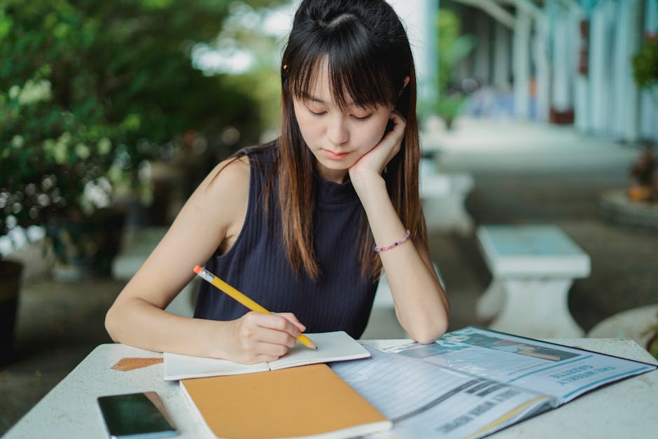 Concentrated young Asian female student sitting at table and writing in copybook while doing home task